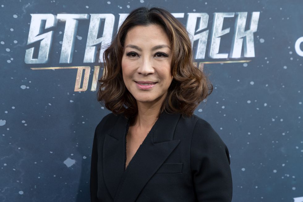 Michelle Yeoh zambind, purtand o camasa neagra, stand in fata unui fundal cu stele si cuvintele Star Trek Discovery pe ele