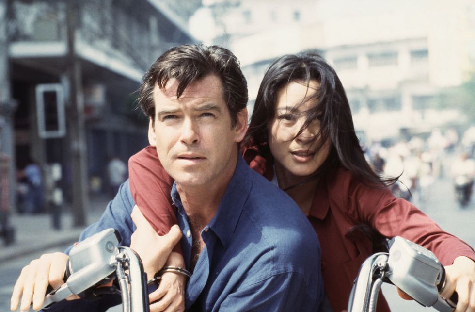 Pierce Brosnan purtand o camasa albastra si Michelle Yeoh purtand o camasa rosie, stand pe o motocicleta pe un drum de oras