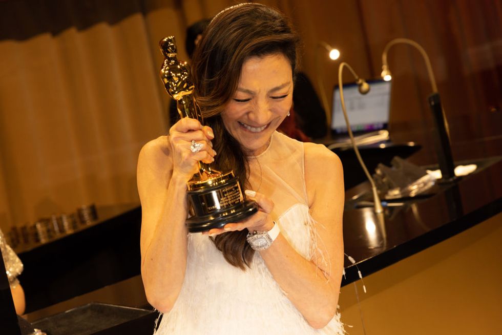 Michelle Yeoh, zambind si privind in jos cu ochii inchisi, tinand un premiu al Academiei, purtand o rochie alba si stand in fata unei mese intr-o camera intunecata